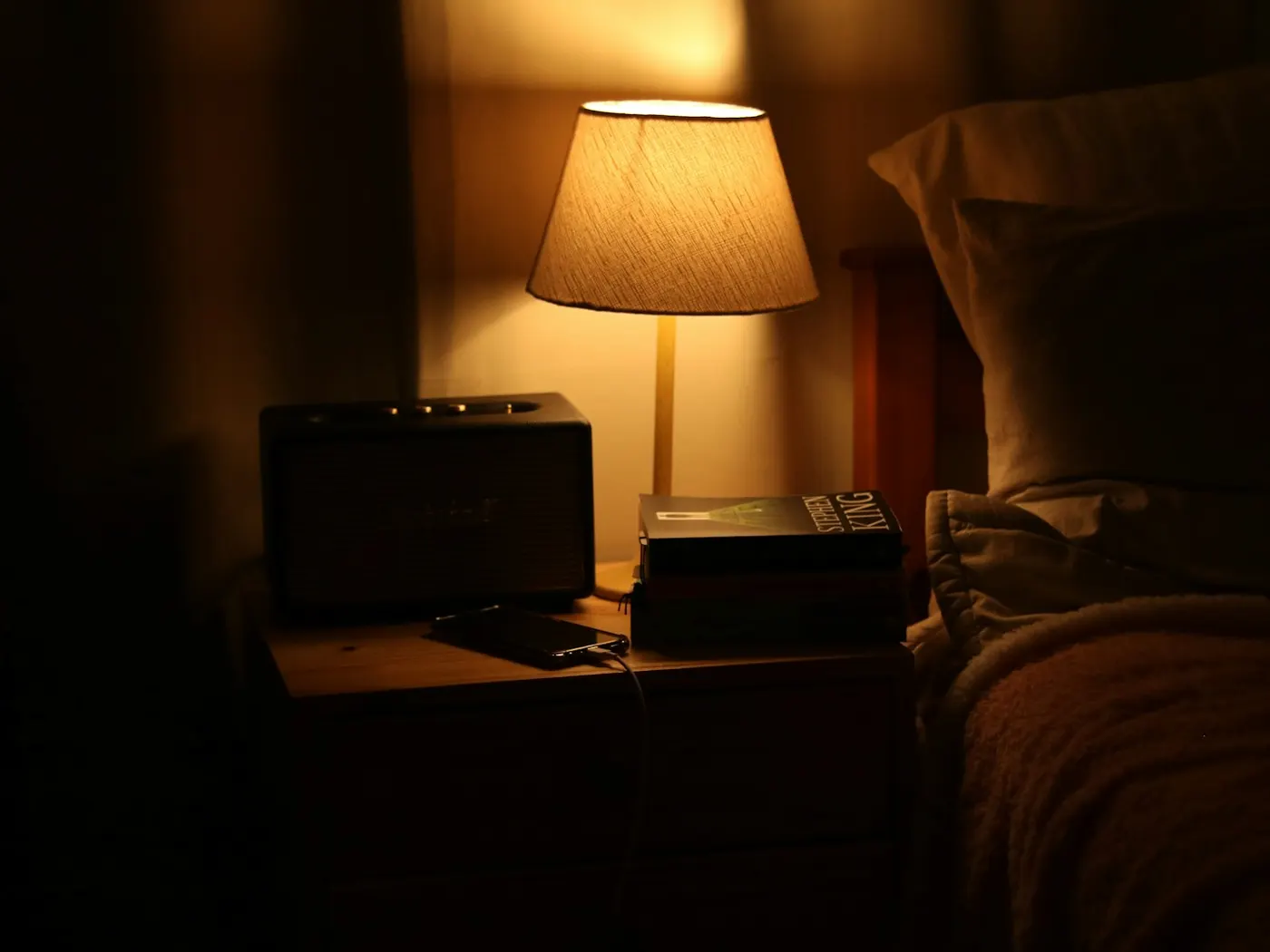A bedside table at night under a lamp, with a small device beside the bed